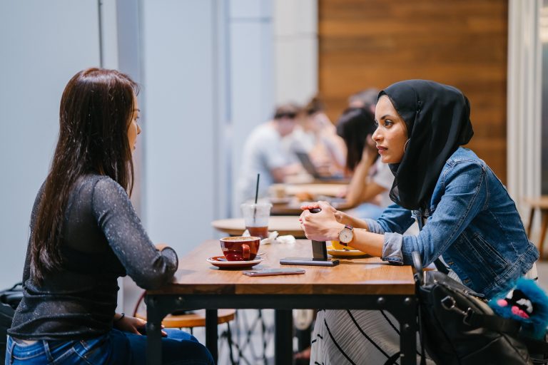 Two women having a conversation at a cafe table, one wearing a hijab.