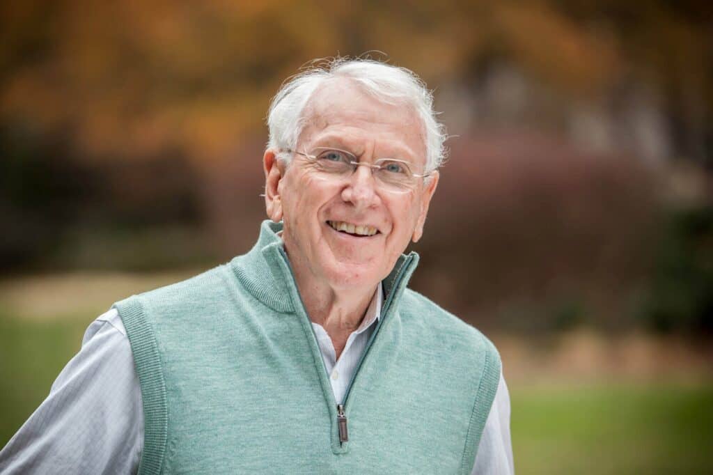 A man smiling outdoors in a park during autumn, wearing glasses and a light green vest.