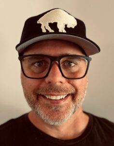 Smiling man wearing glasses and a black cap with a bear logo.