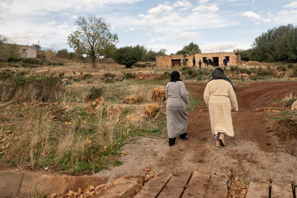 Two women walking on a rural path towards a small village in a dry landscape.